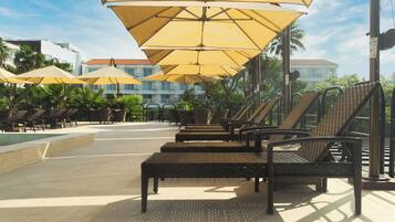 A poolside area with sun loungers and umbrellas, a tiled floor, and buildings in the background.