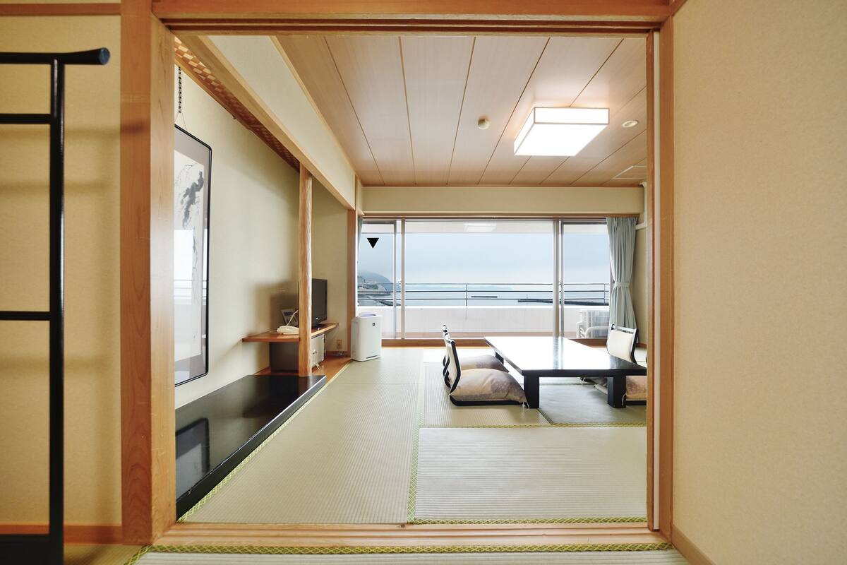 A traditional Japanese room with tatami flooring, a futon bed, a television, and a view of the sea.