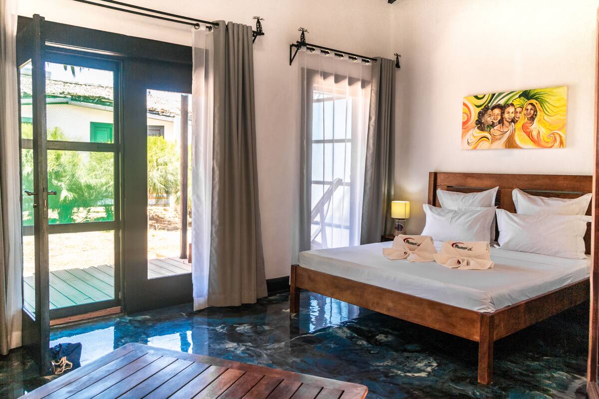 A hotel room with a bed, a wooden table, and a view of the outdoors through a glass door.