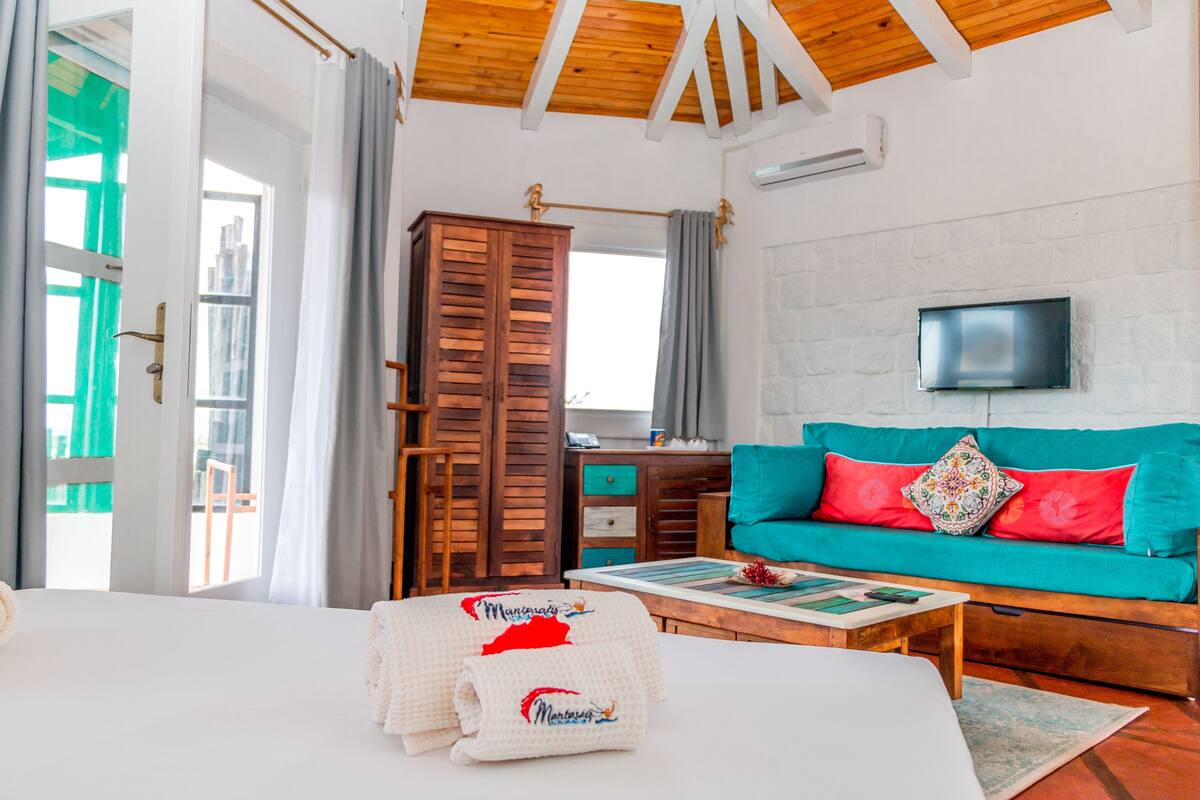A hotel room with a bed, a wooden wardrobe, a teal sofa with red cushions, a television mounted on the wall, and a window with curtains.