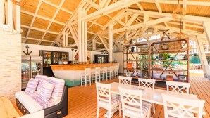 Un amplio espacio al aire libre con suelo de madera, mobiliario blanco y una barra con distintos artículos.