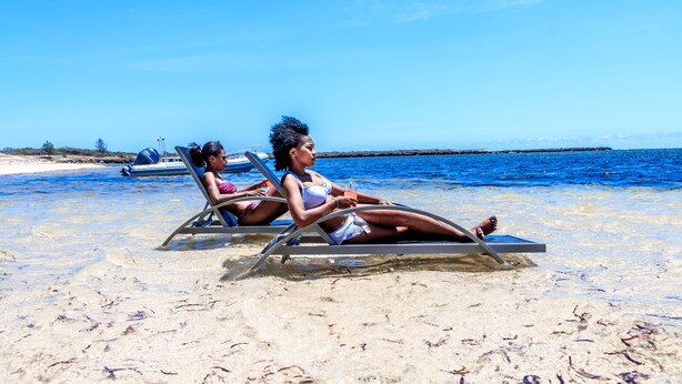 Dos personas descansando en sillas de playa en una arena clara, con agua azul transparente y un barco al fondo.