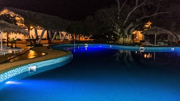 Une piscine éclairée pour la baignade nocturne, bordée de carreaux, entourée de chaises longues et de parasols, avec des arbres en arrière-plan.