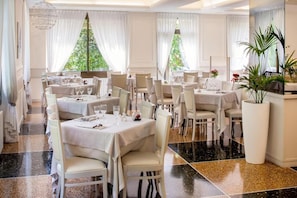 A restaurant with multiple tables, white tablecloths, and chairs, set in a room with large windows and a plant in the corner.