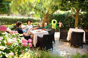 An outdoor dining area with wicker furniture and stone flooring, surrounded by lush greenery and flowering plants.