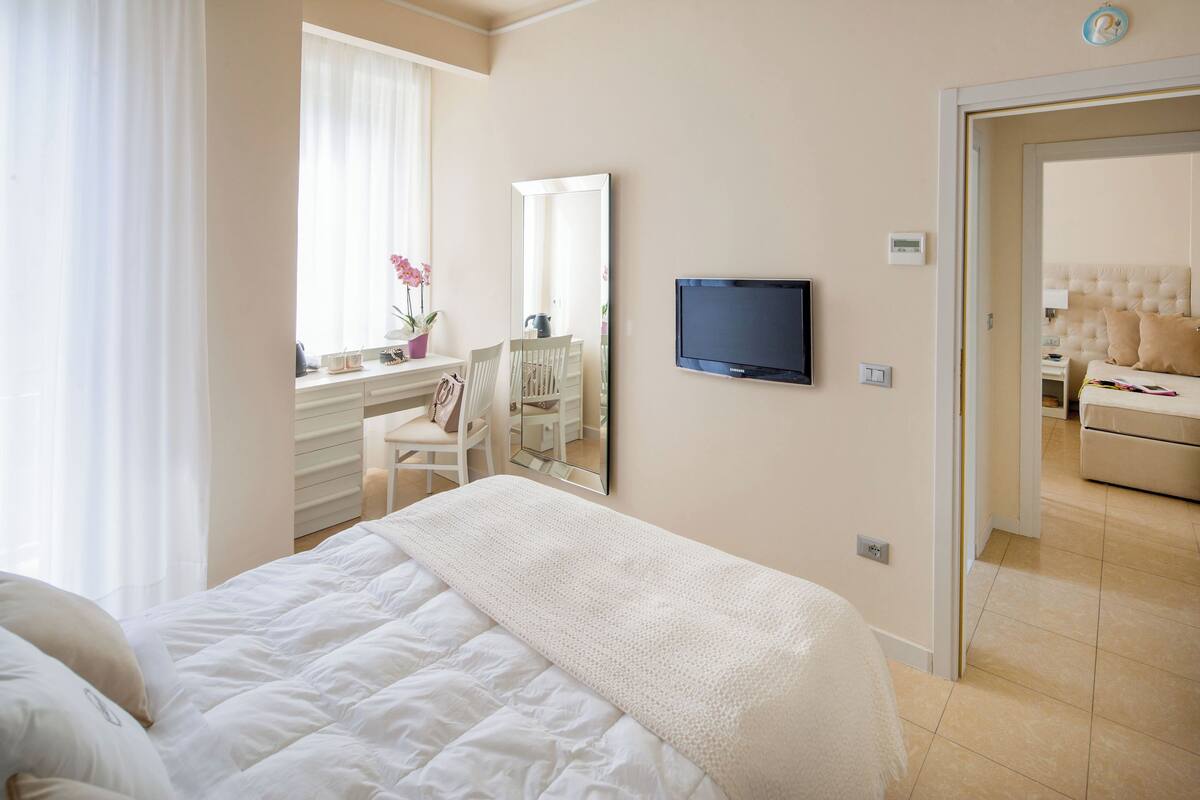 Een hotelkamer met een groot bed, een televisie, een spiegel, een bureau met een stoel, en uitzicht op een andere kamer.
