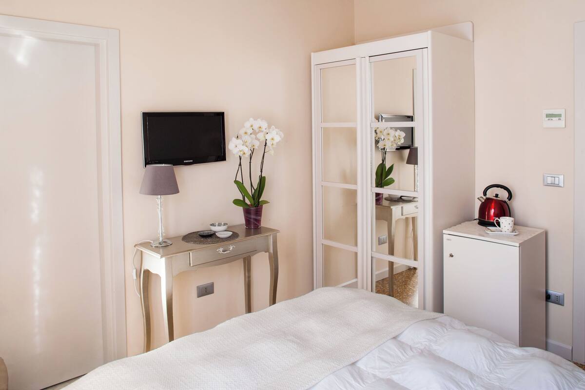 Hotelkamer met een bed, bureau met lamp, televisie, kast en een klein kastje met waterkoker en kop.