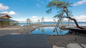 Eine Holzterrasse führt zu einem Kiesstrand mit einem kleinen Teich, umgeben von Bäumen und blauem Himmel.