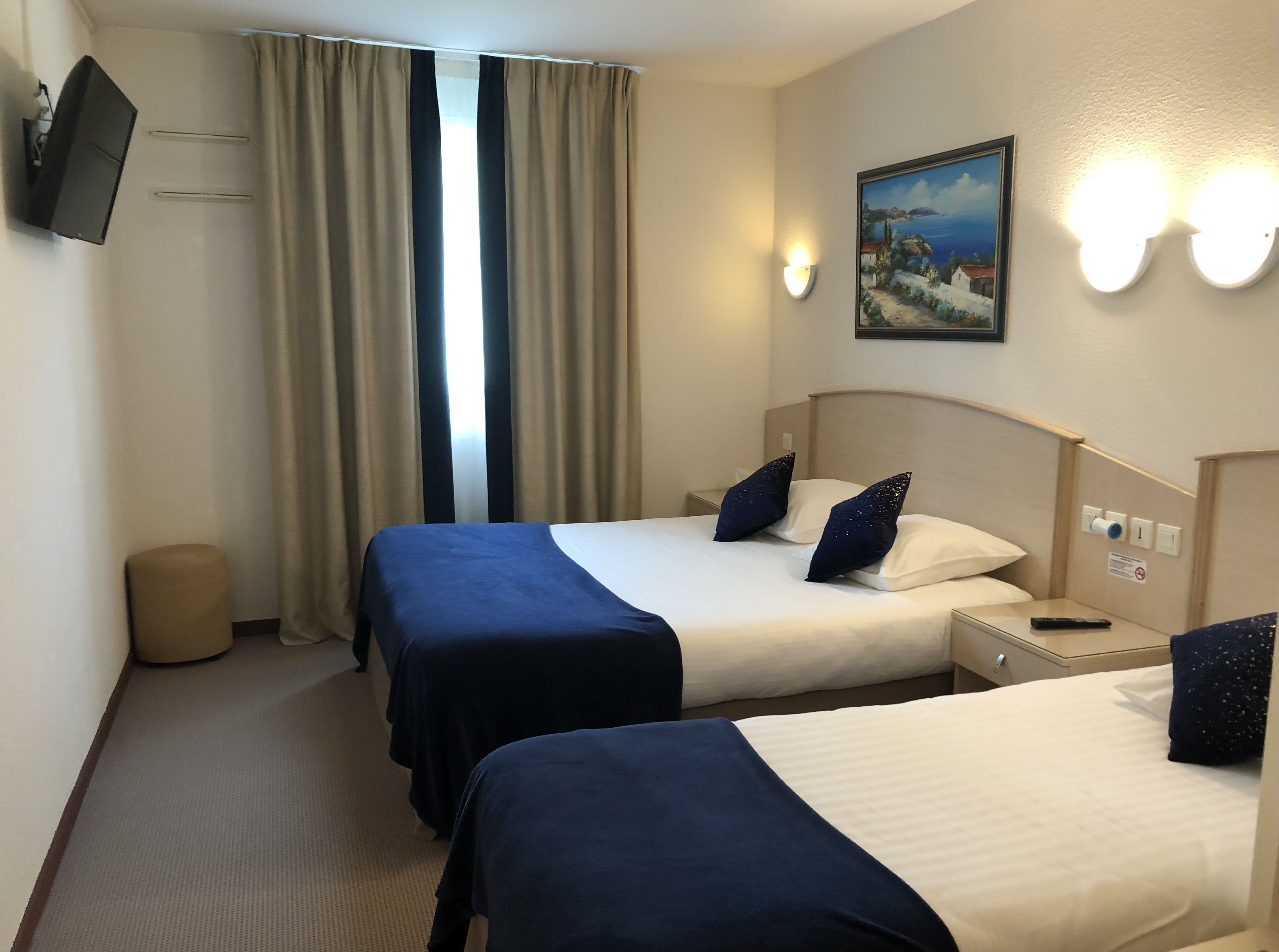 Ein Hotelzimmer mit zwei Betten, einem an der Wand montierten Fernseher, einem Gemälde und einem Nachttisch mit Fernbedienung.