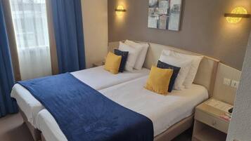 A double bed with a blue blanket, two bedside tables, a wall-mounted abstract painting, and a window with curtains.