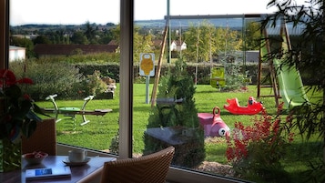 Vista desde una habitación con un ventanal grande que da a un jardín con un columpio, tobogán y un estanque.