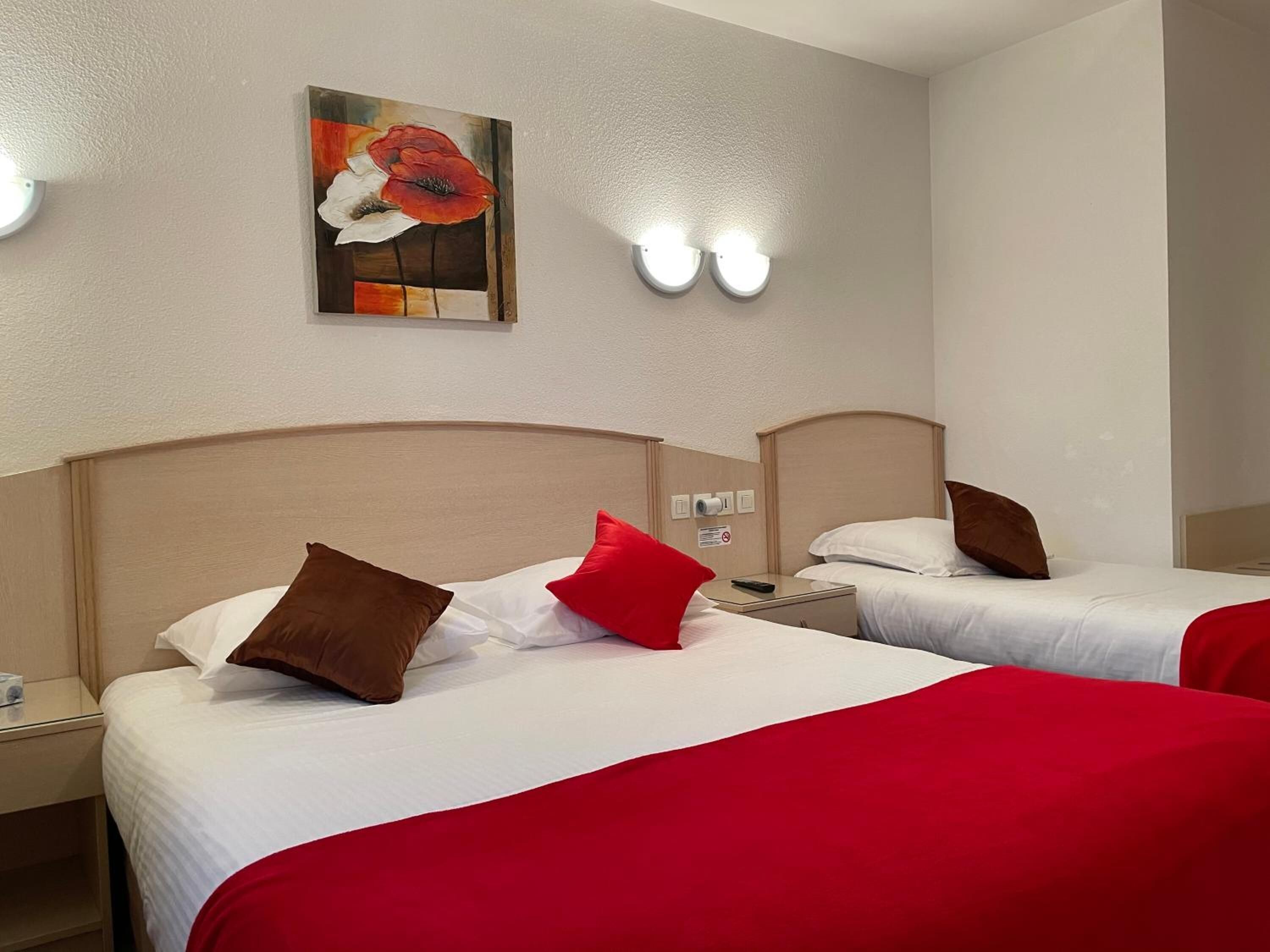 A hotel room with two beds, each with white bedding and red accents. There is a painting on the wall above the beds, and a nightstand with a remote control and a lamp.