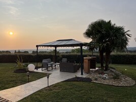Un área de estar al aire libre con techo, con sillas y una mesa, rodeada por un jardín con una palmera y un mueble de madera decorativo.