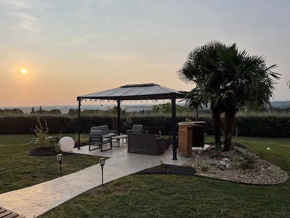 Un área de estar al aire libre con techo, con sillas y una mesa, rodeada por un jardín con una palmera y un mueble de madera decorativo.