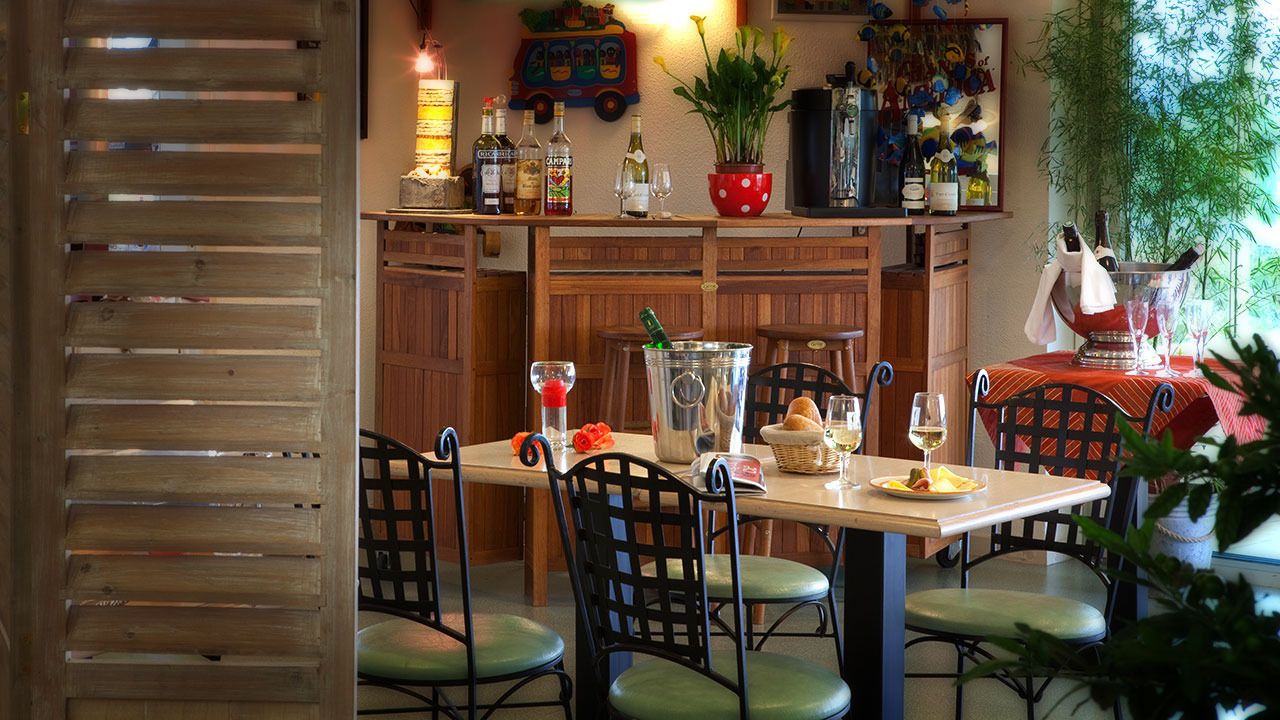 A dining area with a wooden table, black chairs, and a red tablecloth. There are bottles and glasses on the table, and a potted plant in the background.