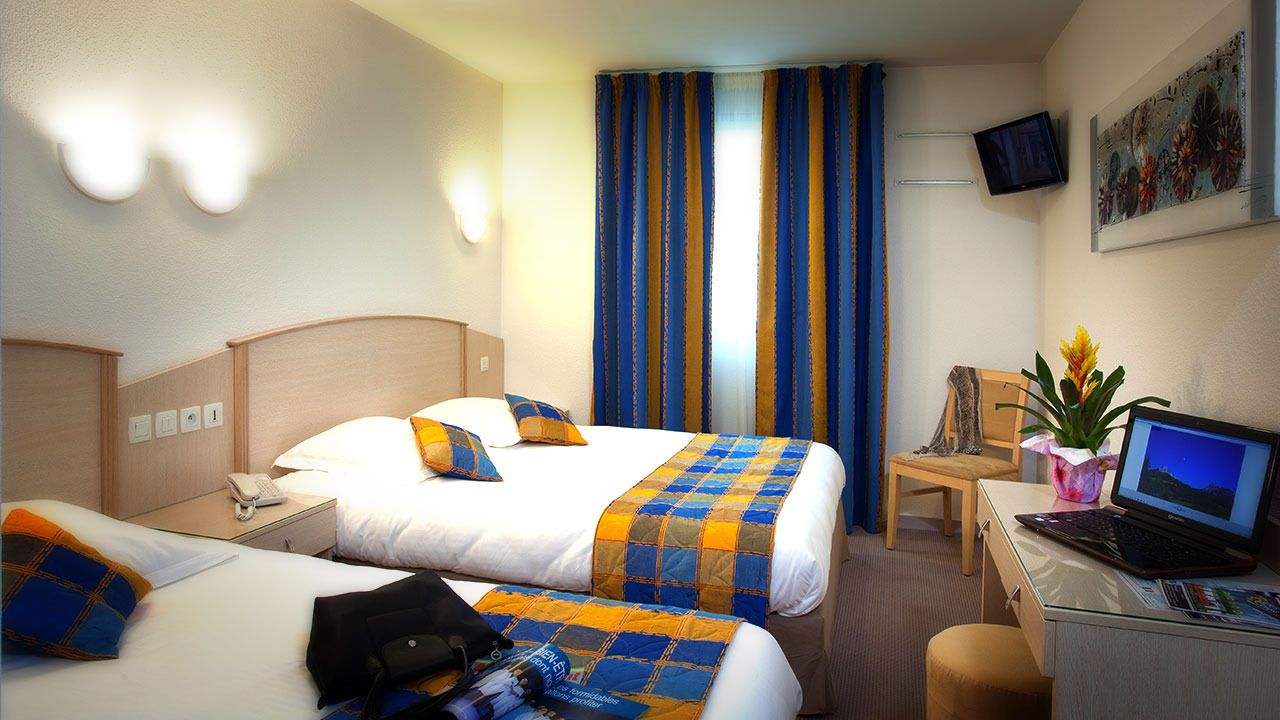 Une chambre d’hôtel avec deux lits, un bureau avec un ordinateur portable, une chaise, un téléviseur fixé au mur et une fenêtre avec des rideaux.
