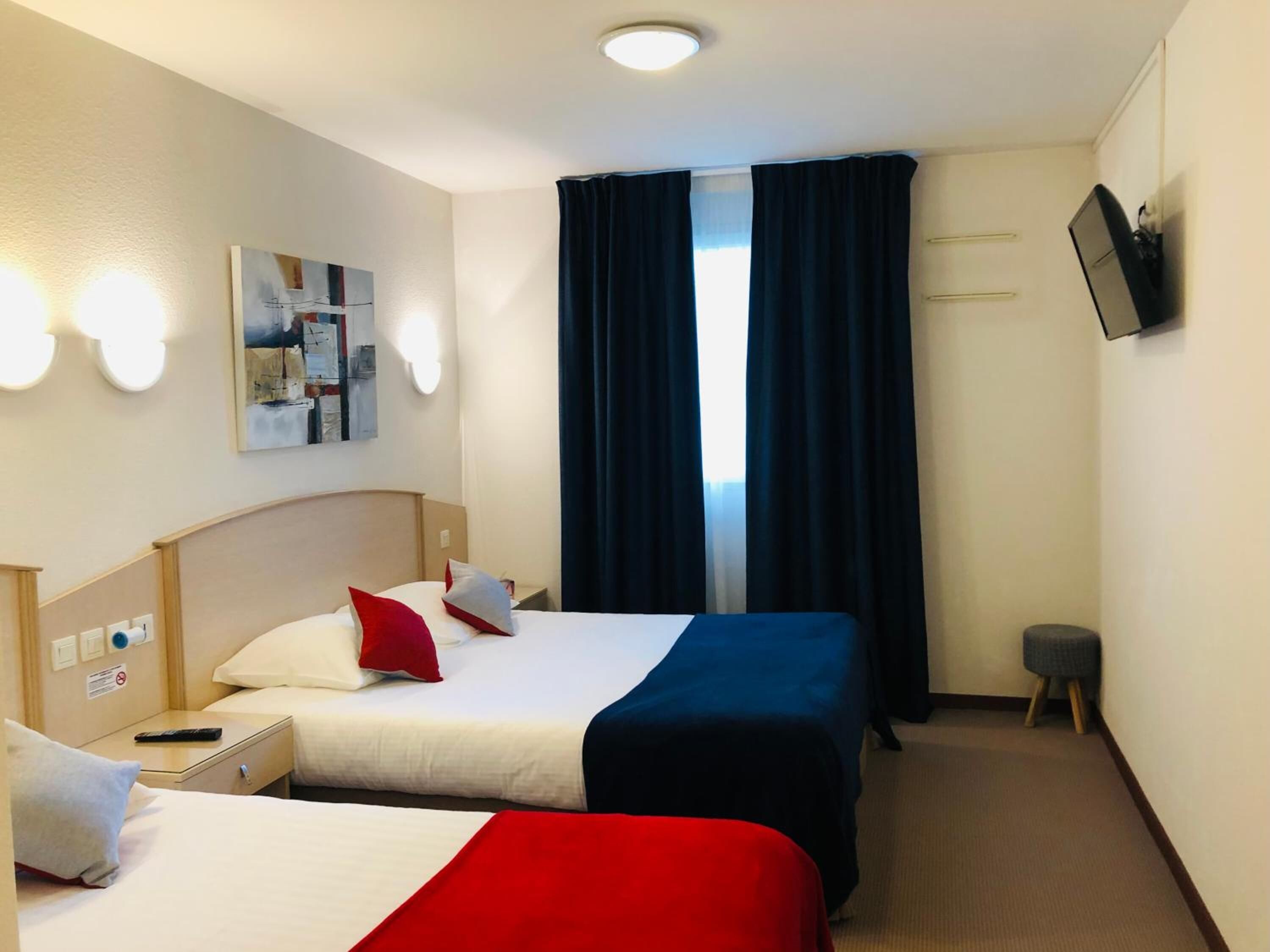 Habitación de hotel con dos camas, un televisor de pantalla plana montado en la pared, un cuadro sobre las camas y una ventana con cortinas oscuras.