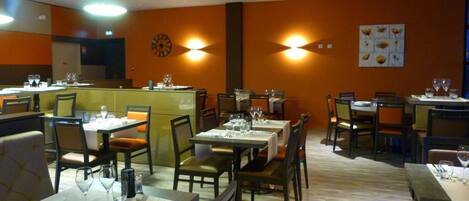 A restaurant with multiple tables and chairs, a central counter, and wall-mounted lighting.