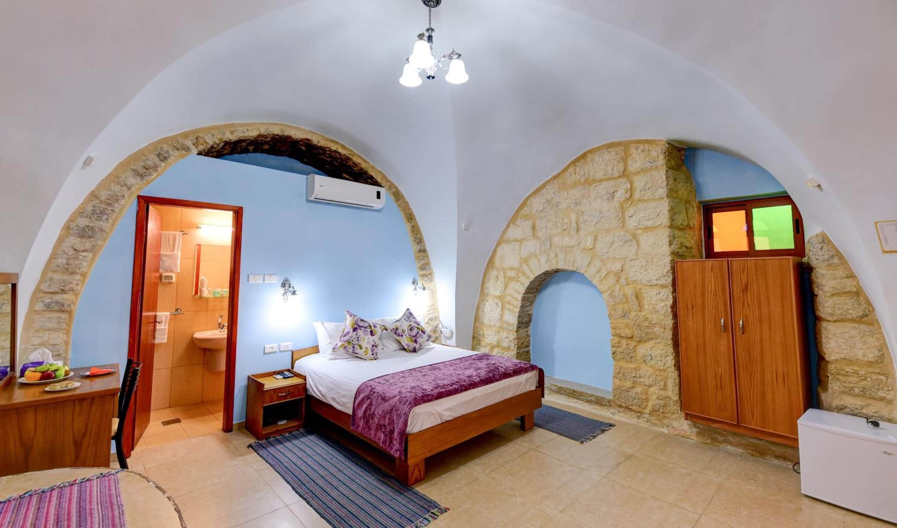 Ein Schlafzimmer mit Bett, einem Holzschrank, einem Schreibtisch mit Obst und einem durch einen Bogen gewölbten Durchgang sichtbaren Badezimmer.