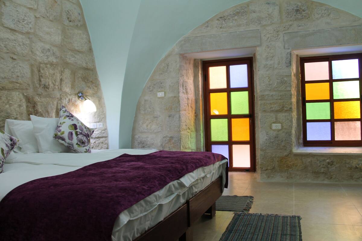 A bedroom with a stone wall, a bed with a purple blanket, and a window with colorful panes.