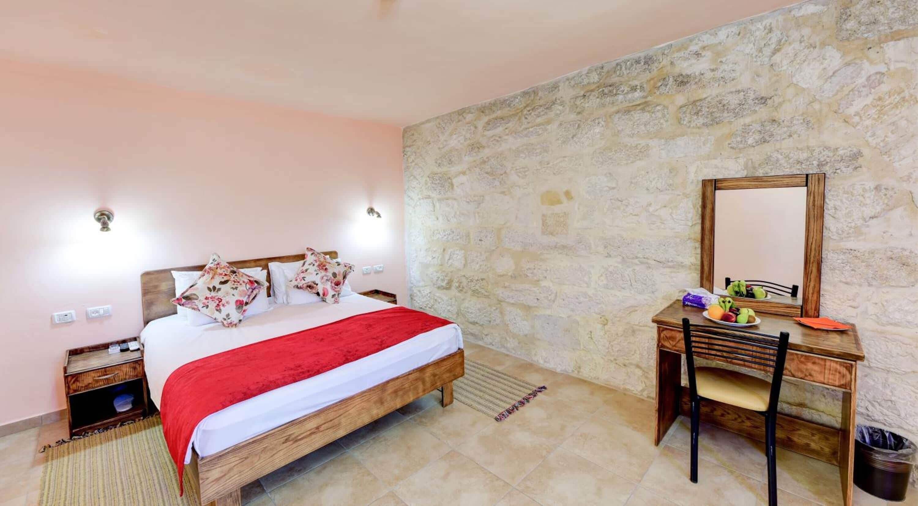 A bedroom with a wooden bed, a desk with a chair, a mirror, and a stone wall.