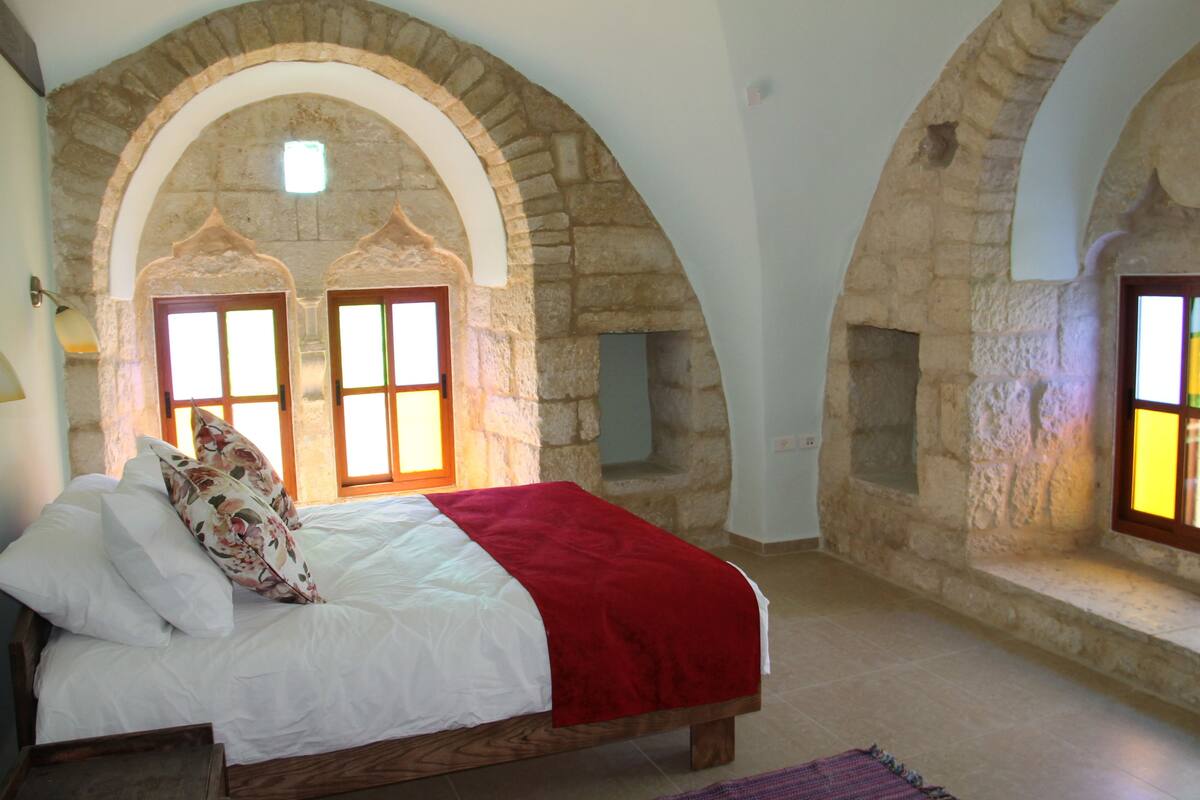 A bedroom with a bed, floral pillows, a red blanket, and stone arches.