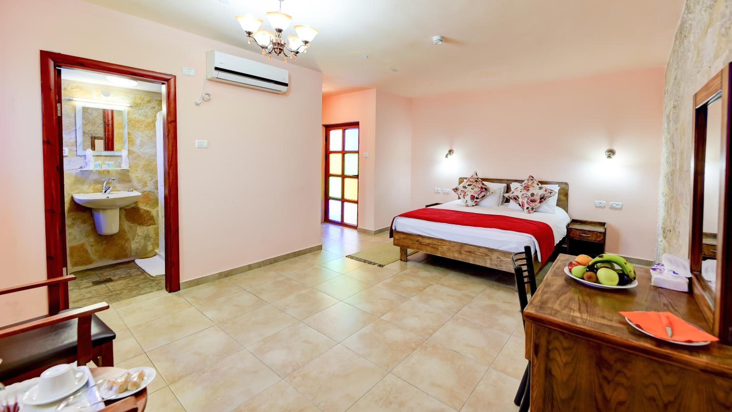 Una habitación de hotel con una cama, un escritorio con una silla, una pequeña mesa con frutas, un espejo y un equipo de aire acondicionado.