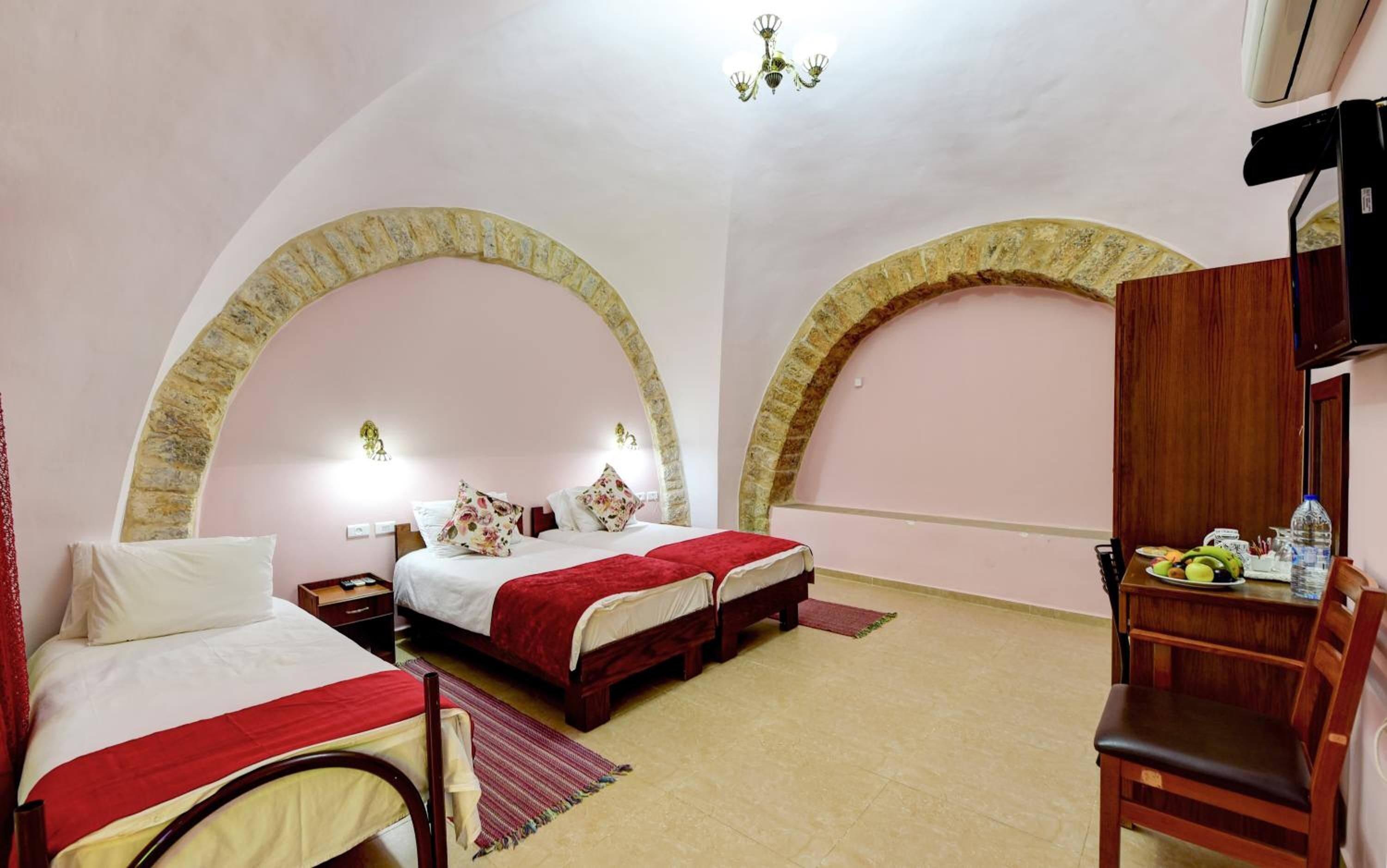 A hotel room with two beds, a wooden wardrobe, a desk with a chair, and a chandelier.