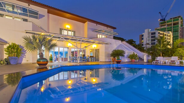 A swimming pool with a covered patio area, potted plants, and a view of a building with multiple floors and a construction crane in the background. - Sharrow Bay (Pattaya)