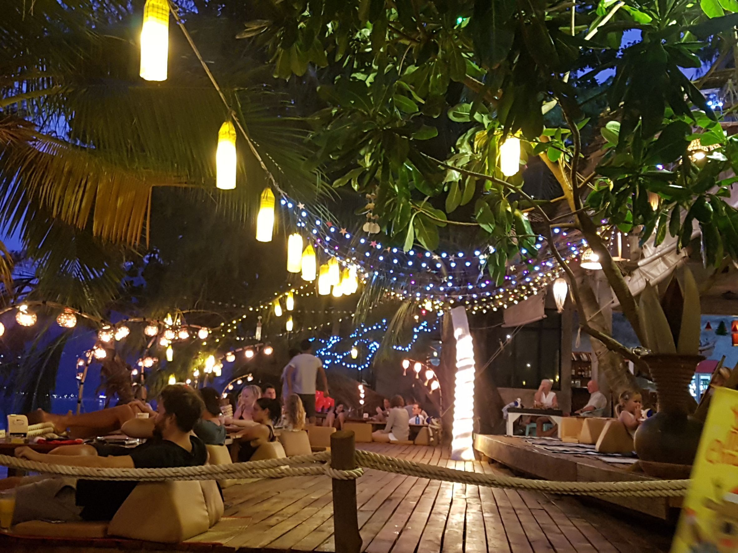 An outdoor seating area with tables and chairs, surrounded by trees and string lights.