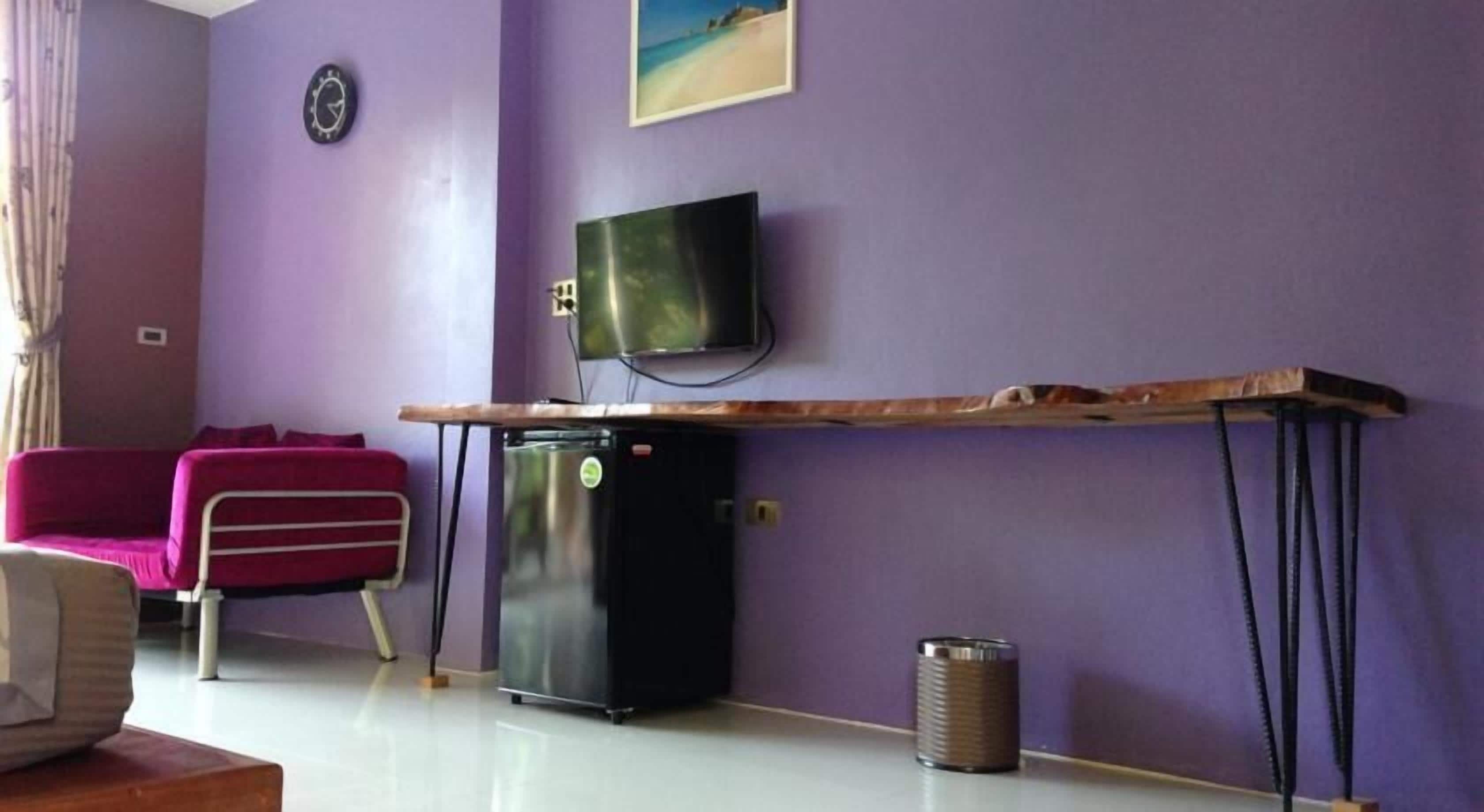 Una habitación con pared morada, un televisor de pantalla plana sobre una mesa de madera, un refrigerador negro pequeño y un sofá rosa, además de un cubo de basura.