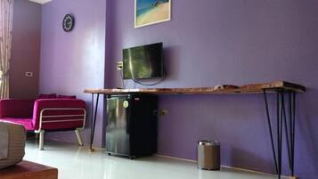 Una habitación con pared morada, un televisor de pantalla plana sobre una mesa de madera, un refrigerador negro pequeño y un sofá rosa, además de un cubo de basura.