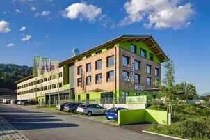 Un hotel moderno con fachada verde y marrón, múltiples ventanas y un estacionamiento con varios coches.
