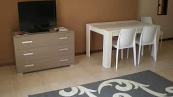 A room with a beige dresser, a television on top, a white table with four chairs, and a patterned rug on the floor.