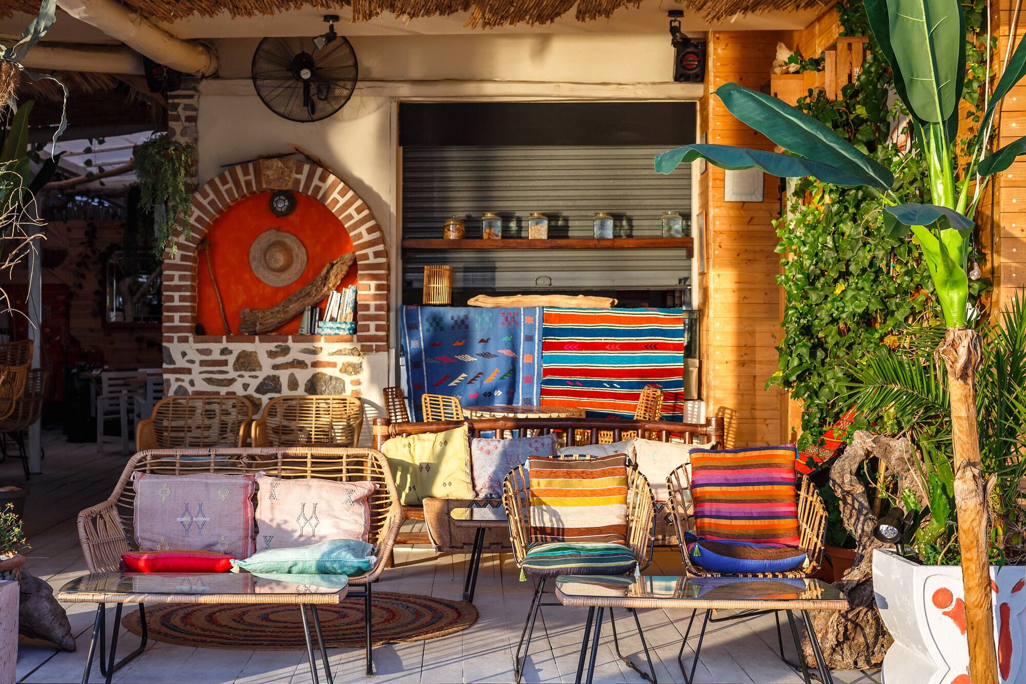 Un espacio de estar al aire libre con sillas y cojines de mimbre, techo de paja y diversos elementos decorativos, incluyendo una gran obra de arte circular en la pared y textiles coloridos.