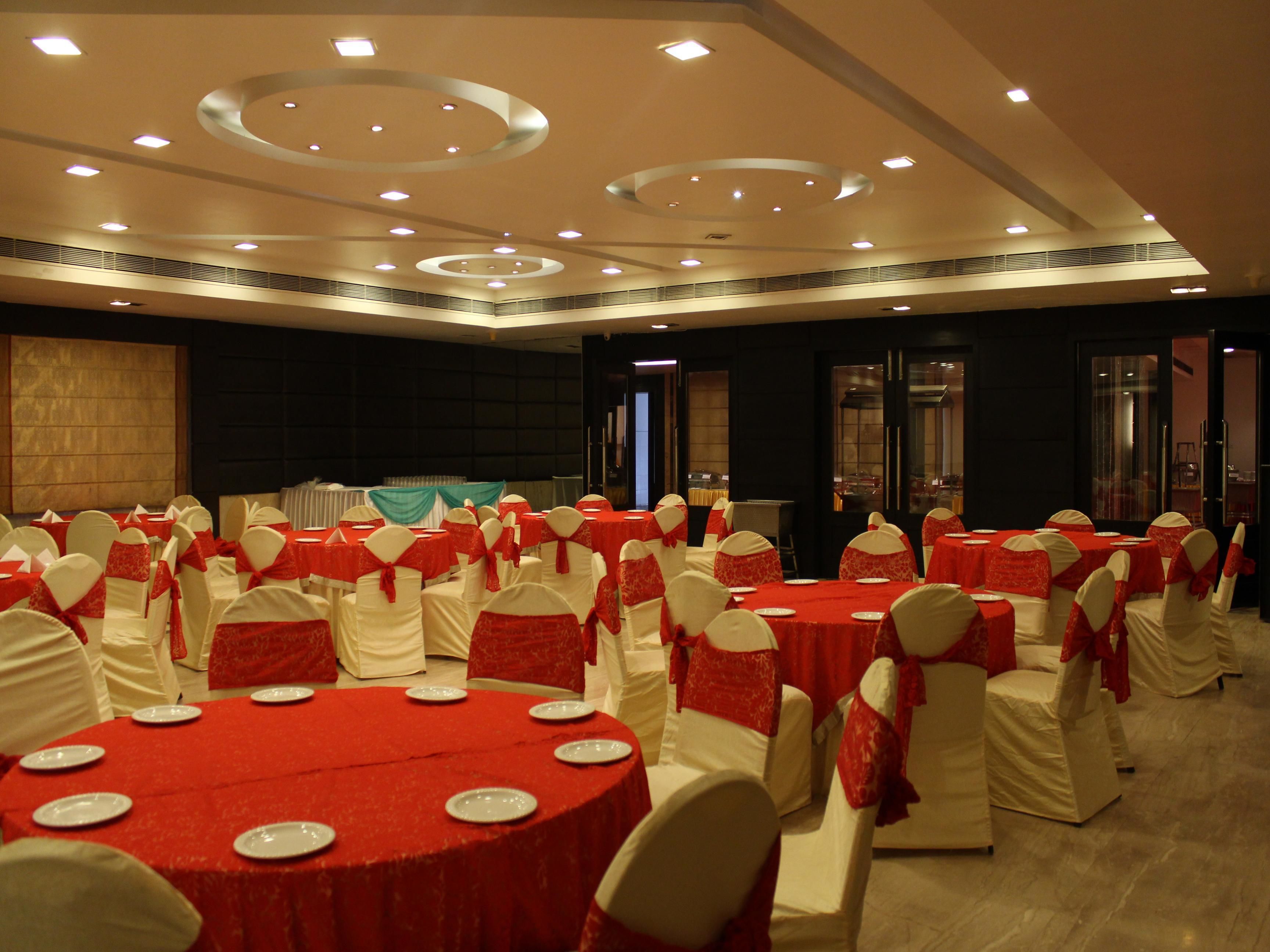 A banquet hall with round tables covered in red tablecloths, white chairs with red sashes, and recessed lighting on the ceiling.