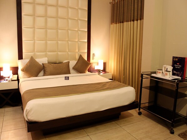 A hotel room with a large bed, a nightstand with a lamp, a small table with a coffee maker, and curtains.