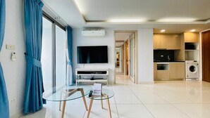 A modern living room with a glass coffee table, a flat-screen TV, and a kitchen area with a washing machine. - Laguna Bay 2 by PSR ASIA (Pattaya)