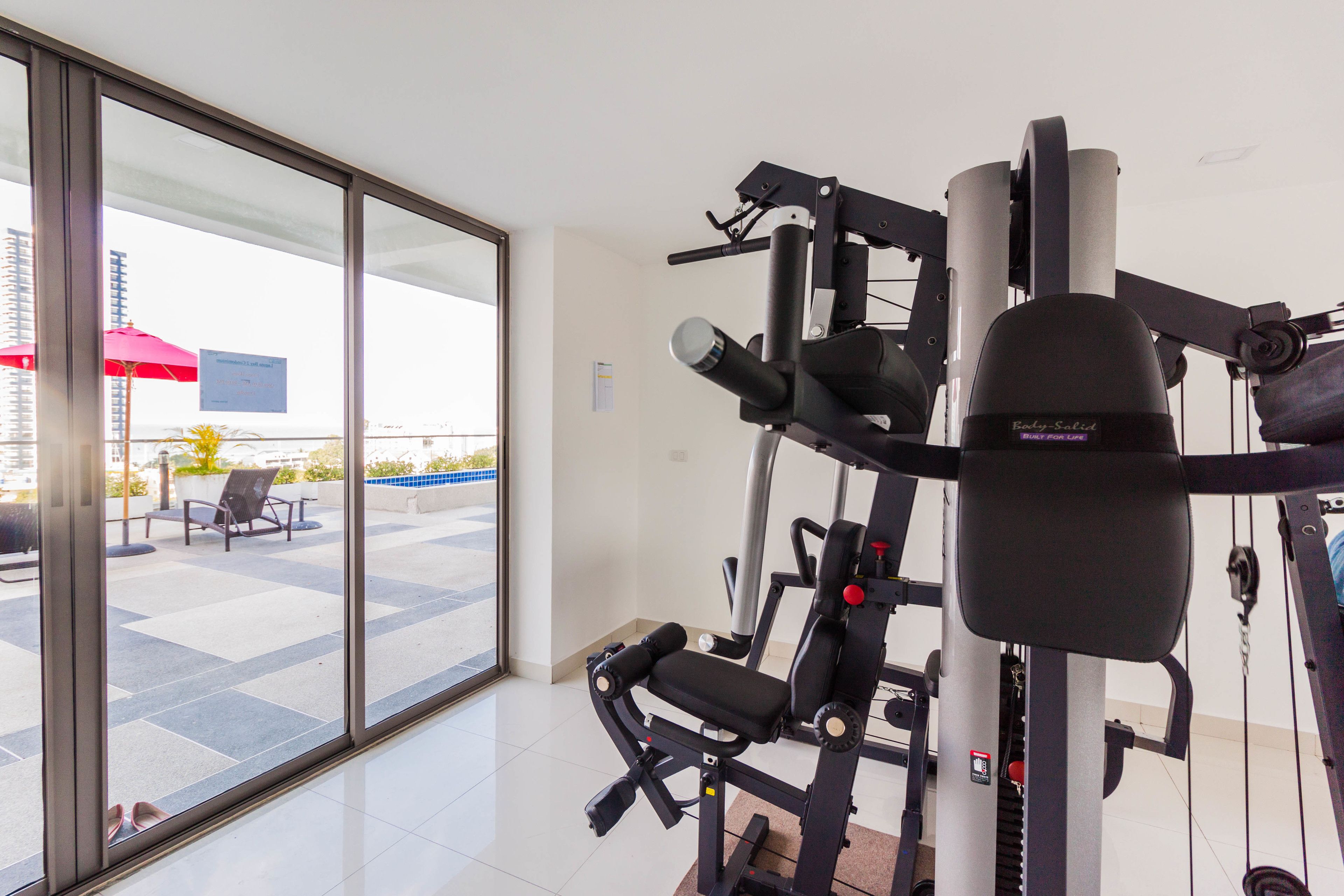 Una palestra con attrezzature per esercizi e vista su un patio esterno con sedie e un ombrellone rosso.