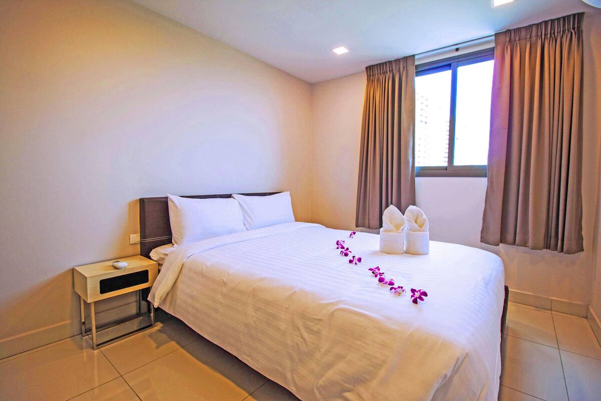 A hotel room with a large bed, a nightstand, a window with curtains, and a view of the outside.