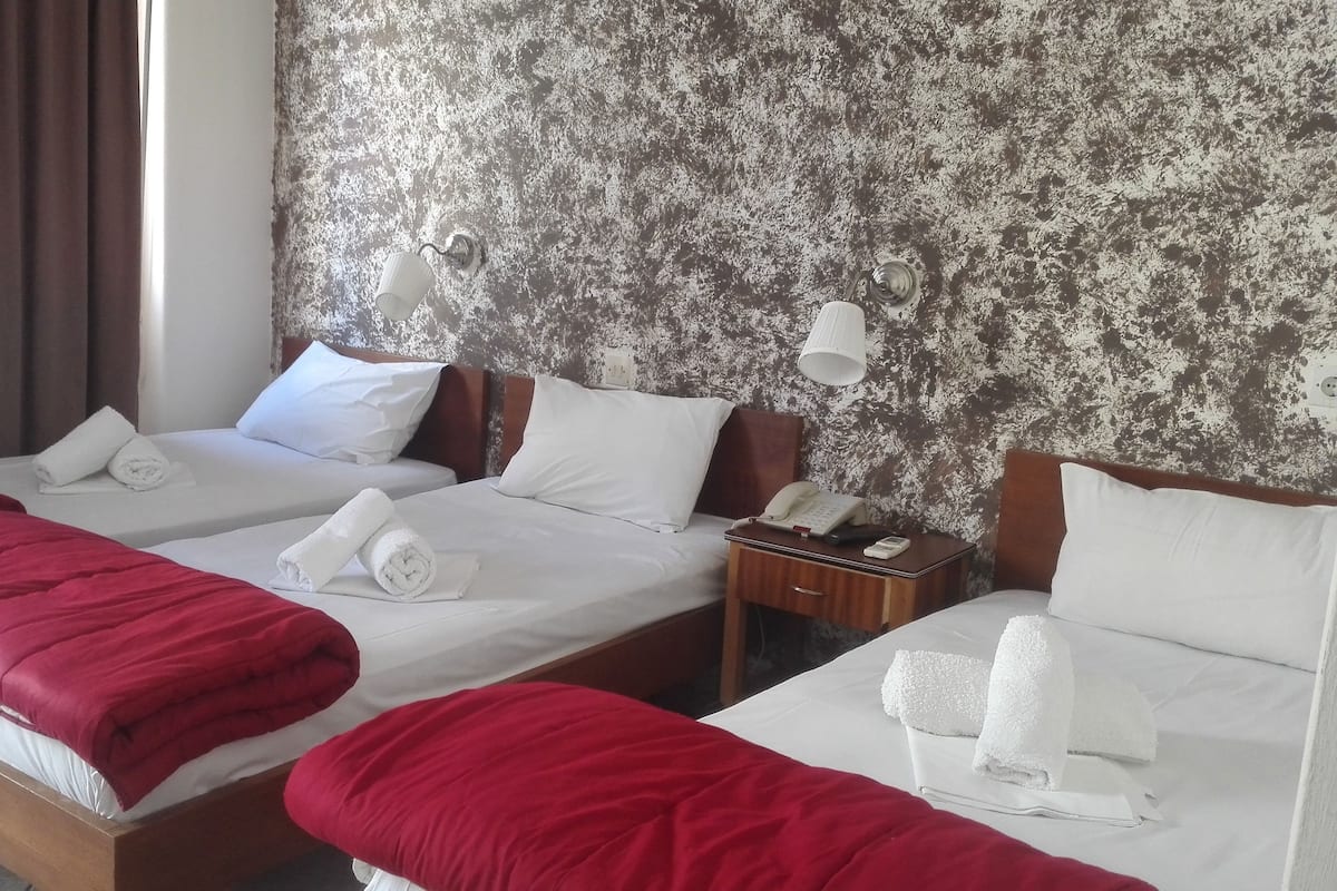 Habitación de hotel con dos camas, cada una con sábanas blancas y mantas rojas. Hay dos mesitas de noche, una con una lámpara y la otra con un teléfono y una mesita pequeña. La habitación tiene una pared con textura y una ventana con cortinas.