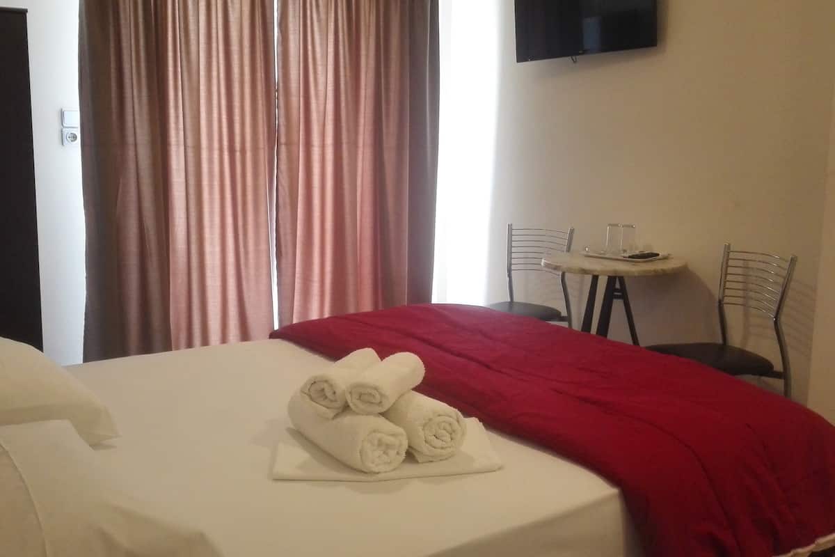 Una habitación de hotel con cama, una mesa con sillas, un televisor montado en la pared y un ventanal con cortinas.