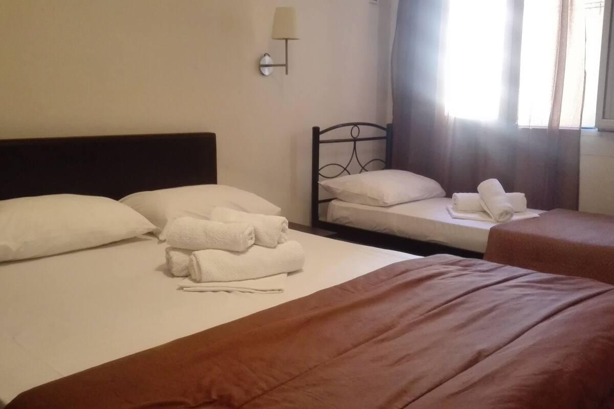 Habitación de hotel con dos camas, cada una con sábanas y almohadas blancas, una mesita de noche, una lámpara de pared y un ventanal con cortinas.