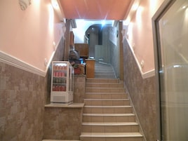 Una escalera con una máquina expendedora a la izquierda, que lleva a un espacio con pared de azulejos y una puerta a la derecha.