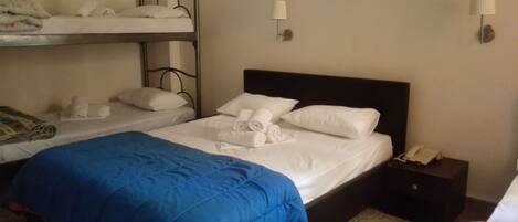 A hotel room with two beds, one with a blue blanket and the other with white bedding. There are towels on the beds, a nightstand with a phone, and a wall-mounted lamp.