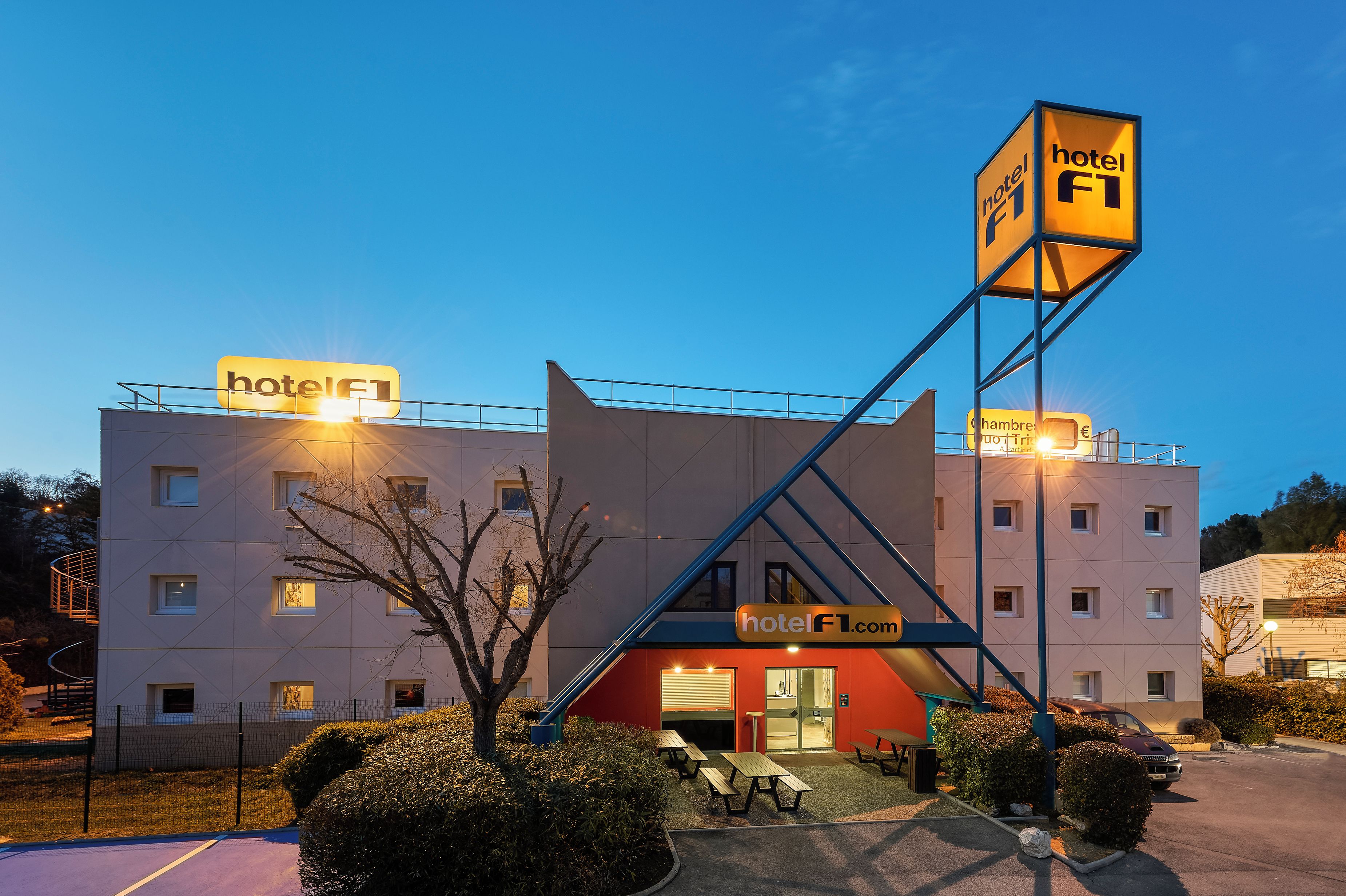 Een hotel met een opvallend 'hotel F1' bord, een rode ingang en een parkeerplaats met picknicktafels.