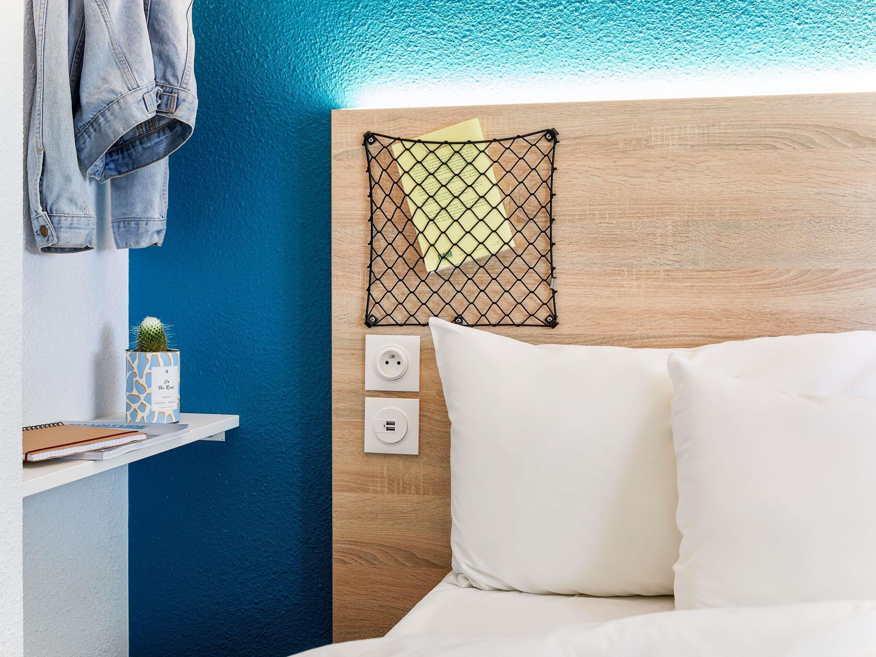 A neatly arranged hotel room with a wooden headboard, a small cactus on a shelf, a bed with white linens, and a wall-mounted power outlet.