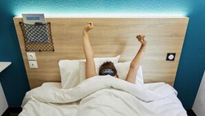 Een persoon ligt in bed met armen omhoog, een klein netje op de hoofdbord en een stopcontact aan de muur.