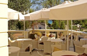 Une terrasse extérieure avec des meubles en osier blanc et des parasols, entourée de verdure et d’arbres.