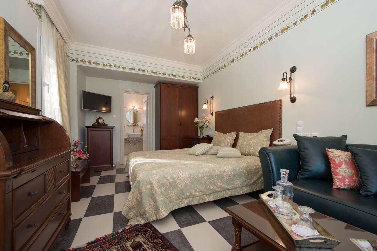Une chambre d’hôtel avec un sol à carreaux, un lit recouvert d’une literie à motifs floraux, un canapé en cuir, une commode en bois et un téléviseur.